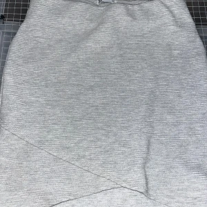 Ljusgrå ribbad kortkjol XS - Snygg ljusgrå ribbad kortkjol från C i storlek XS. Kjolen har en enkel och stilren design med omlott-detalj framtill. Perfekt för dig som gillar en clean och modern look. Superenkel att matcha med både sneakers och boots.