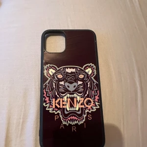 Kenzo mobilskal med tigertryck  - Snyggt mobilskal från Kenzo med ikoniskt tigertryck i lila, rosa och beige toner på mörk bakgrund. Skalet har Kenzo Paris-loggan framtill och är gjort i tålig syntet. Passar dig som vill ha en cool och unik look till mobilen.