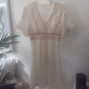 Cremefärgad,  med beige spets. Bohostil. - Superfin Cremefärgad midiklänning med blommig spets och v-ringad hals. Klänningen har tofsar framtill, korta ärmar med spetskant och broderade detaljer. Perfekt för sommarens alla tillfällen, med romantisk och bohemisk vibe.