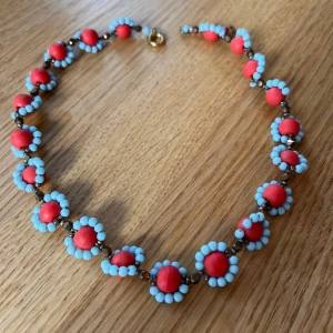 Färgglatt halsband - Unikt halsband med stora röda pärlor omgivna av små ljusblå pärlor. Pärlorna är sammanlänkade med bruna, blänkande detaljer och halsbandet har ett guldfärgat lås. Välgjort. Längd ca 42 cm. Inköpt i antikaffär. 