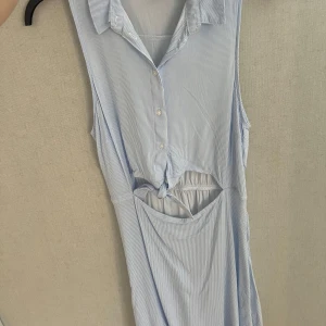 Blåvit randig klänning H&M - Säljer en fräsch blåvit randig klänning från H&M DIVIDED i storlek 40. Klänningen är ärmlös med krage, knappar framtill och har en snygg omlott-detalj med knyt i midjan och cut-out. Perfekt för varma dagar och har en lätt, luftig känsla.