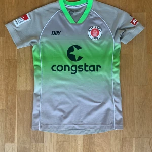St. Pauli grå/grön fotbollströja XS - Snygg St. Pauli fotbollströja i unisex XS med grå bas och ljusgröna detaljer. Tröjan har korta ärmar, V-ringad krage med grön kant och tryckta loggor från Congstar och St. Pauli. Tillverkad av återvunnet material, perfekt för match eller träning.