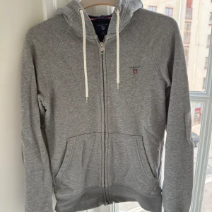 Grå hoodie från GANT med dragkedja - Snygg grå hoodie från GANT med vit snörning i huvan och praktisk dragkedja framtill. Tröjan har två stora fickor och diskret GANT-logga på bröstet. Tillverkad i mjuk bomull och polyesterblandning, perfekt för chill dagar. Långärmad och med huva för extra komfort.