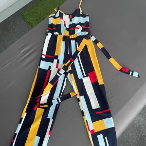 Färgglad jumpsuit med grafiskt mönster - Säljer en snygg jumpsuit med grafiskt mönster i blått, gult, rött, vitt och svart. Jumpsuiten har smala axelband, markerad midja med knytband och raka ben. Perfekt för dig som gillar färg och vill sticka ut.
