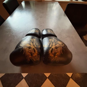 Svarta boxningshandskar från Joya - Säljer ett par svarta boxningshandskar från Joya med snyggt guldigt drakmotiv på ovansidan. Handskarna har bred kardborrestängning och är tillverkade i syntetmaterial. Perfekta för dig som vill sticka ut på träningen!