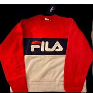 Röd och vit Fila sweatshirt - Snygg sweatshirt från Fila i rött, vitt och marinblått med stor logga framtill. Tröjan har lång ärm och klassisk passform, perfekt för en sportig och avslappnad stil. Tillverkad i mjuk bomullsmix som känns skön mot huden.
