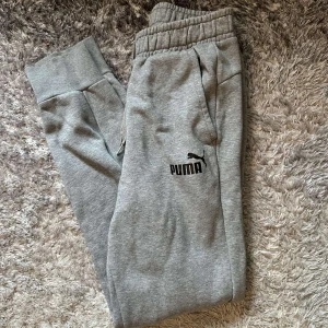 Grå mjukisbyxor från Puma - Snygga grå mjukisbyxor från Puma med elastisk midja och dragsko. Byxorna har ribbade muddar vid bensluten, två sidofickor och Puma-logga tryckt på låret. Perfekta för chill eller träning, tillverkade i mjuk bomullsblandning.