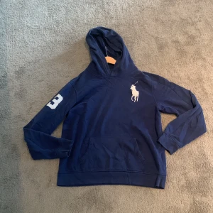 Marinblå hoodie från Polo Ralph Lauren - Snygg marinblå hoodie från Polo Ralph Lauren med stor vit logga på bröstet och siffran 3 på ärmen. Tröjan har huva, känguruficka och långa ärmar. Perfekt för chill dagar eller när du vill ha en sportig look.det är XL i kids men är samma som S