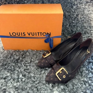 Louis Vuitton klackskor  - Snygga klackar från Louis Vuitton i mörkbrunt med klassiskt monogrammönster. Skorna har spetsig tå, låg klack och guldfärgade spännen med logotyp. Materialet är canvas med detaljer i skinn. Perfekt för dig som vill sticka ut med lyxig designerstil.