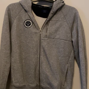 Grå hoodie GANT x Båstad S - Grå hoodie från GANT x Båstad med dragkedja, mörkblå huva och bröstficka med zip. Snyggt tennisinspirerat märke på bröstet. Perfekt för dig som gillar sportig stil och vill ha något bekvämt och stilrent. Passar dig som vill ha en klassisk hoodie med modern touch. Ursprungligt pris: 1300-1900kr.