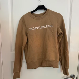 Beige sweatshirt från Calvin Klein Jeans - Snygg beige sweatshirt från Calvin Klein Jeans med vit logga framtill. Tröjan har rund halsringning, långa ärmar och ribbade muddar vid ärmslut och nederkant. Perfekt för en chill och stilren look.