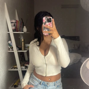 Vit croppad ribbad zip-top - Säljer en vit croppad topp med långa ärmar och dragkedja framtill. Toppen är ribbad och har en tight passform som framhäver figuren. Perfekt att styla med jeans eller kjol för en trendig look.