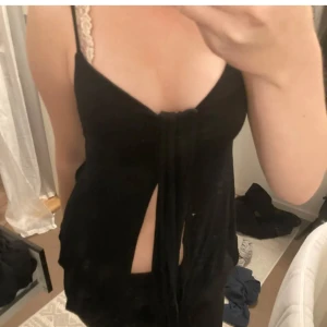 Svart linne med slits och knyt - Snygg svart linne med smala axelband och djup v-ringning. Framtill finns en cool slits och knytning som ger en trendig look. Perfekt för dig som gillar att sticka ut och vill ha något unikt till garderoben.