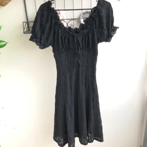 Svart klänning med puffärm - Supersöt svart kortklänning i ett blommigt mönster tyg med puffiga små kortärmar, som går att sätta som off-shoulder och söt snörning vid halsringningen. Den är mycket stretch i så den passar från S till L