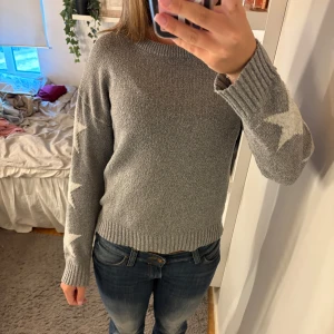 Grå stickad tröja med stjärnor - Mysig grå stickad tröja med vita stjärnor längs ärmarna. Tröjan har rund halsringning, ribbade muddar och en relaxed fit. Perfekt för dig som gillar en enkel men ändå cool look med lite extra detaljer på ärmarna.