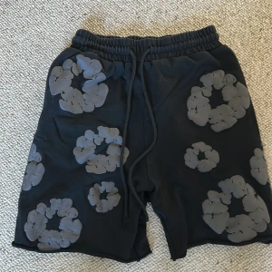 Svarta shorts med grått blommönster - Unika svarta shorts med gråa blommiga mönster över hela tyget. De har elastisk midja med snörning och en bakficka. Perfekta för dig som gillar streetwear och vill sticka ut med cool design. 
