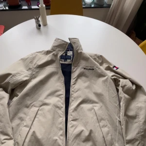 Beige vindjacka från Tommy Hilfiger - Snygg beige vindjacka från Tommy Hilfiger med klassisk ståkrage, dragkedja och två fickor framtill. Jackan har logga broderad på bröstet och flagga på ärmen. Perfekt för vår och höst, lätt att matcha och riktigt clean stil. Sitter som storlek S.