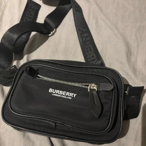 Svart axelväska från Burberry - Säljer en svart axelväska från Burberry med justerbar rem där loggan är invävd. Väskan har flera fack, dragkedja framtill och detaljer i skinn. Perfekt för att bära det viktigaste och ger en clean, lyxig vibe.