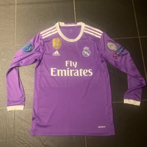Real Madrid Ronaldo 7 lila långärmad tröja - Säljer en lila långärmad Real Madrid fotbollströja från Adidas med Ronaldo 7 på ryggen. Tröjan har vita detaljer, Champions League 2016 patch på ärmen, FIFA-märke och Fly Emirates-tryck på bröstet. Tillverkad i lätt och ventilerande climacool-material.