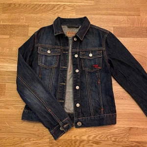 Jeansjacka - Snygg jeansjacka från Zoul i mörkblå denim med kontrasterande sömmar och silverfärgade knappar. Jackan har två bröstfickor med lock och en röd broderad detalj på ena fickan. Knappt använd!