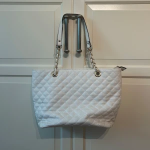 Vit quiltad axelväska med kedjedetaljer - Säljer en stilren vit axelväska med quiltat mönster och kedjedetaljer på axelbanden. Väskan har dragkedja baktill och rymligt huvudfack. Perfekt för dig som vill ha en klassisk och trendig accessoar.