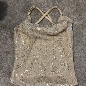 Glittrig silverfärgad paljett-topp med smala axelband - Säljer en snygg glittrig topp i silver med paljetter och smala korsade axelband. Perfekt för att sticka ut på festen! Toppens material är syntet och den är ärmlös med croppad passform.