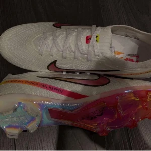 Nike Air Zoom Mercurial Vapor 15 Elite FG Marcus Rashford X Disruption Pack - White/Black/Flash Crimson/Pink - Säljer ett par vita Nike fotbollsskor i syntetmaterial med coola regnbågsdetaljer och inspirerande text på sidan. Skorna har snörning och en färgglad sula som verkligen sticker ut. Perfekta för att göra ett statement på planen!