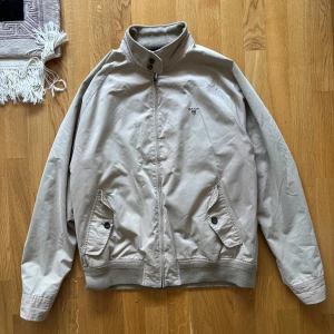 Beige jacka från Gant - Klassisk beige jacka från Gant med hög krage och dragkedja framtill. Jackan har två fickor med knappar och ribbade muddar vid ärmslut och nederkant. Fodret är rutigt i rött, blått och vitt. Perfekt för en stilren look.