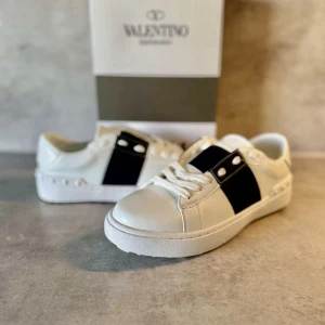 Vita sneakers Valentino Garavani med svart detalj - Säljer ett par vita Valentino Garavani sneakers med bred svart rem och silverfärgade nitar på sidorna. Skorna har rund tå, platt sula och är tillverkade i skinn. Klassisk och stilren design med tydlig logga på plösen.