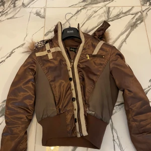  Baby Phat jacka - Säljer en brun bomberjacka från Baby Phat med pälskant på huvan och broderad logga på ryggen. Jackan har dragkedja, knappar och ribbade muddar. Detaljer i beige och guld samt en ficka med dragkedja på bröstet❤️ Den är egentligen XL men passar mig som XS!! 