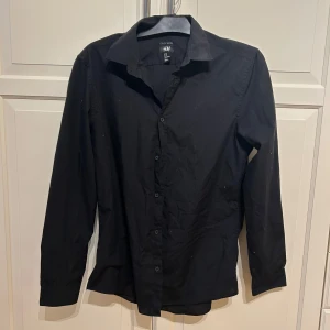 Svart skjorta från H&M - Klassisk svart skjorta från H&M med lång ärm och knappar framtill. Skjortan har en stilren design med krage och är tillverkad i ett lättskött material som är easy iron. Perfekt för en clean och enkel look. I strl M