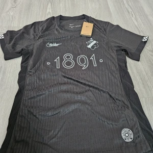 Svart AIK fotbollströja från Nike - Säljer en svart AIK fotbollströja från Nike med trycket '1891' på bröstet och AIK:s klubbmärke. Tröjan är kortärmad och har diskreta ränder i tyget samt märkning för 'Dri-FIT ADV'. Perfekt för dig som vill visa ditt stöd för AIK.