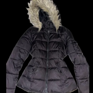 Svart pufferjacka med snatched waist - Svart pufferjacka med luva. Jackan har dragkedja framtill och två fickor med knappar. Perfekt för kalla dagar när du vill hålla dig varm och snygg. Den har även en snached passform som gör att kroppen får en hourglass figur