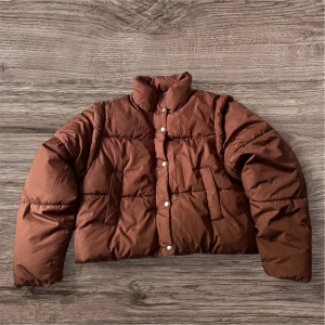 Brun pufferjacka från Gina Tricot - Säljer en brun pufferjacka från Gina Tricot med hög krage och tryckknappar framtill. Jackan är croppad och har två fickor på sidorna. Perfekt för kyligare dagar när du vill ha en stilren look.