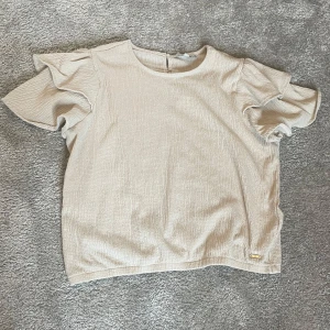 Beige blus med volangärmar  - Säljer en beige blus från Cream med korta volangärmar och rund halsringning. Blusen har en liten knappdetalj i nacken.