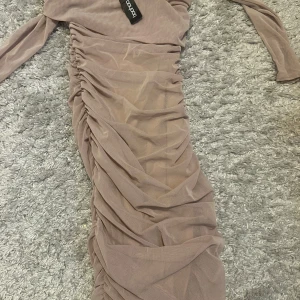 Beige långklänning från Boohoo - Säljer en beige långklänning från Boohoo med lång ärm och snygg rynkad design längs hela klänningen. Materialet är mjukt och stretchigt, perfekt för dig som vill ha en klänning med lite extra detaljer. Aldrig använt. Har en i storlek 34 och en i storlek 36. 