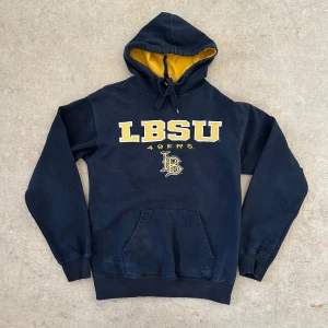 Svart LBSU hoodie med gul text - Säljer en svart hoodie från LBSU med gul text och logga framtill. Tröjan har huva med gul insida och dragsko samt en stor magficka. Perfekt för dig som gillar college-stil!                                      Vintage college hoodie  LBSU 49ers   Size S   The hoodie is great condition   Length:65cm Chest:50cm Arm-length:58cm