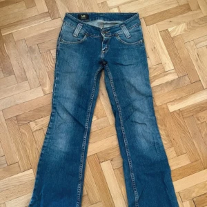 Blå bootcut jeans från Lee - Säljer ett par klassiska blå bootcut jeans från Lee med snygga slitningar och femficksmodell. Jeansen har normal passform och är tillverkade i bomull med lite stretch för extra komfort. Perfekta till vardags för en avslappnad look. Bra skick förutom en fläck på fickan