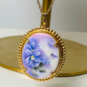 Vintage halsband med handmålat blommotiv - Unikt halsband med en oval berlock i guldtonad metall och handmålat motiv av lila och blå blommor med gröna blad. Kedjan är tunn och elegant, perfekt för att ge en romantisk touch till din stil.