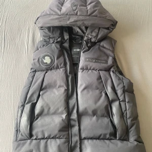 Grå dunväst från Sail Racing - Säljer en grå dunväst från Sail Racing med huva och dragkedja. Västen har två stora fickor framtill och en snygg logotyp på bröstet. Perfekt för lager-på-lager och stilren look.