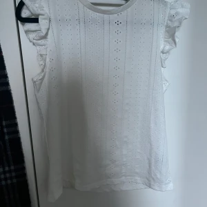Vit ärmlös blus med volang från SHEIN - Säljer en söt vit ärmlös blus från SHEIN med broderade detaljer och volang vid axlarna. Perfekt för vår och sommar, lätt och luftig med fina små hålmönster. Passar till både jeans och kjol!