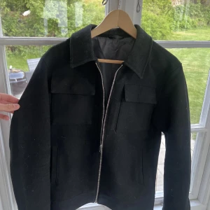 Svart ull overshirt med dragkedja strlk.S - Säljer en stilren svart overshirt med dragkedja framtill och två stora bröstfickor. Jackan har klassisk krage och raka ärmar, perfekt för en clean och modern look under vår/sommar/höst🤩