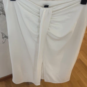Vit kjol med slits framtill.  - Säljer en snygg vit Vit kjol med slits framtill. Kjolen har en elegant drapering och är perfekt för dig som gillar stilrena plagg med en twist. Passar till många olika tillfällen och är enkel att matcha med olika toppar.
