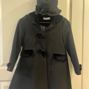 Svart kappa med matchande hatt  - Elegant svart kappa med sammetsdetaljer och rosetter, samt matchande hatt med dekorativ blomma. Kappan har lång ärm, knyt vid ärmslut och fina knappar framtill. Perfekt för kyligare dagar när du vill känna dig extra stilig.