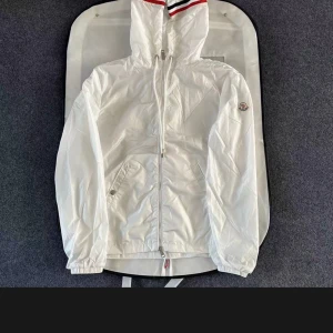 Vit vindjacka från Moncler - Säljer en stilren vit vindjacka från Moncler med huva och dragsko. Jackan har Moncler-logga på ärmen, dragkedja framtill och diskreta knappar med Moncler-gravyr. Röd och marinblå rand på huvan ger en sportig touch.