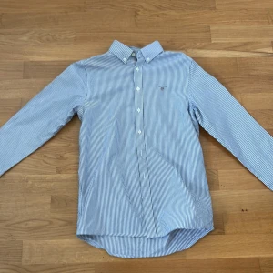 Blårandig skjorta från Gant - Klassisk blårandig skjorta från Gant med button-down krage och broderad logga på bröstet. Skjortan har långa ärmar och knäppning framtill. Perfekt för en stilren och tidlös look.
