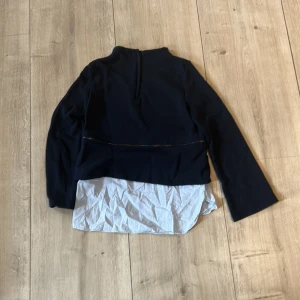 Svart och vit långärmad blus med knyt - Säljer en svart långärmad blus med vit skjortdetalj nedtill och knyt i midjan. Blusen har hög hals och en unik tvådelad look som ger en cool lager-på-lager känsla.