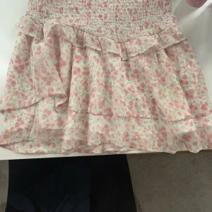 Blommig volangkjol från Gina Tricot - Supersöt kortkjol från Gina Tricot med blommigt mönster i rosa och grönt på vit botten. Kjolen har volanger och smock i midjan för en bekväm passform. Perfekt för vår och sommar!
