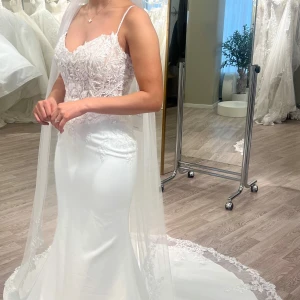 Vit brudklänning med spetsdetaljer och släp - Elegant vit brudklänning med figurnära passform, spetsdetaljer och axelband. Kjolen är slät och lång med ett vackert släp. Klänningen har en romantisk känsla med fina broderier och transparenta partier upptill. Tidlös modell använd en gång. Kemtvättad och vattentvättad. Köpt hos Stockholm brud & fest. 