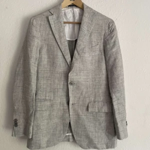 Ljusgrå kavaj från Boggi Milano - Snygg ljusgrå kavaj från Boggi Milano i slim fit-modell. Kavajen har två knappar framtill, klassiska slag och fickor med lock. Tillverkad i ett lätt linnematerial, perfekt för varmare dagar. Helt oanvänd och nypriset ligger på omkring 5 500kr !   Passa på inför sommaren ! 